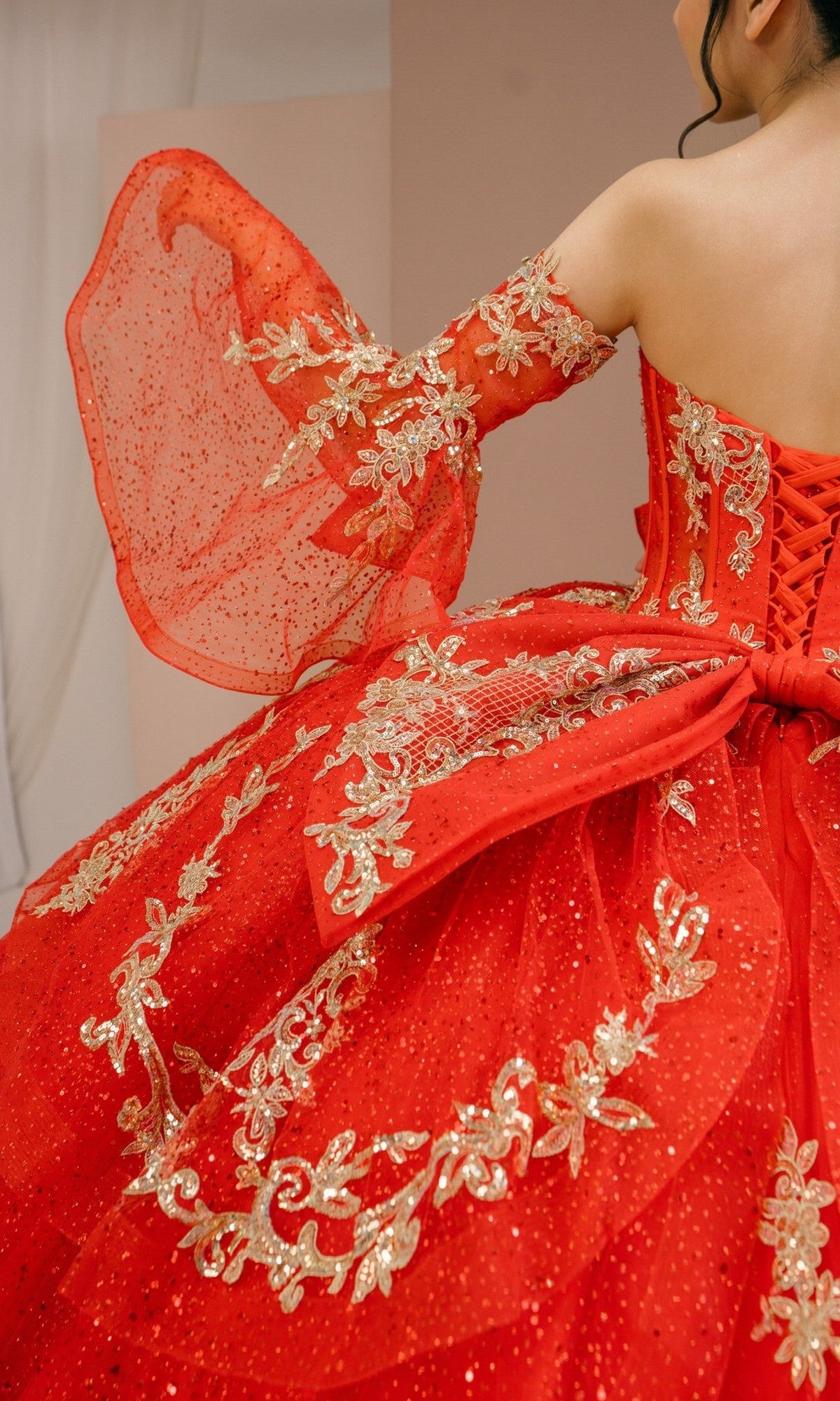 Wearing the Quinceanera Dress 8575JQ by Cinderella Couture, this person is seen from behind in an elaborate red embellished ball gown featuring gold embroidery, glitter tulle, a large bow, and a lace-up back.